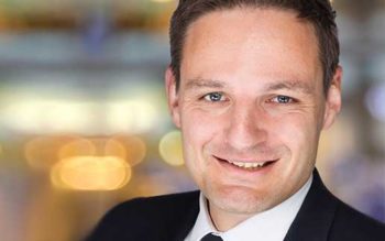 Das Bild zeigt einen Mann in einem Anzug mit einem freundlichen Lächeln. Er hat kurze, dunkle Haare und trägt ein weißes Hemd mit einer dunklen Krawatte. Der Hintergrund ist unscharf und zeigt eine helle, unbestimmte Umgebung, möglicherweise in einem öffentlichen Raum oder einer Veranstaltung.
