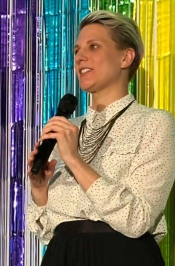 Anja-Kristin Lorke, Managerin für Mitarbeiterbeziehungen DKB Code Factory steht auf einer Bühne und hält ein Mikrofon in der Hand. Sie trägt ein gepunktetes Hemd und eine lange Kette. Im Hintergrund sind farbige, glänzende Vorhänge zu sehen, die eine festliche Atmosphäre schaffen. Sie spricht anscheinend zu einem Publikum über Gleichstellung.