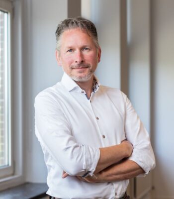 Das Bild zeigt David-Jan Janse, CEO von SurePay - einen Mann mittleren Alters mit einem freundlichen Lächeln. Er trägt ein weißes Hemd mit einem dezenten Muster am Kragen und hat die Arme vor der Brust verschränkt. Sein kurzes, graues Haar und der gepflegte Bart verleihen ihm ein professionelles Erscheinungsbild. Im Hintergrund sind große Fenster zu sehen, die viel Tageslicht hereinlassen und den Raum hell und einladend wirken lassen. Die Wände sind in einem neutralen Farbton gehalten, was den Fokus auf die Person lenkt und eine angenehme Atmosphäre schafft. Der Kontext des Bildes könnte eine Unternehmenspräsentation oder ein professionelles Porträt für eine Website sein. Der Mann strahlt Selbstbewusstsein und Kompetenz aus, was ihn als geeigneten Ansprechpartner in einem geschäftlichen Umfeld erscheinen lässt.