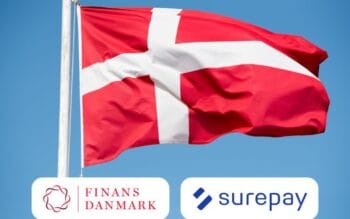 Das Bild zeigt die dänische Flagge, die im Wind weht, begleitet von den Logos von Finance Denmark und SurePay. Finance Denmark, ein Verband für Banken und Finanzinstitute, hat sich für die Implementierung der Verification of Payee (VOP) entschieden, um die Sicherheit im Zahlungsverkehr zu erhöhen. Die Darstellung der Flagge symbolisiert die nationale Identität und den Kontext der Zusammenarbeit im Finanzsektor.