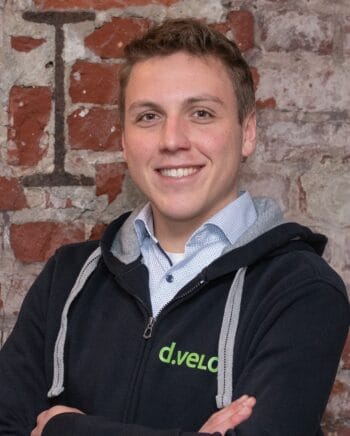 KI darf keine Kredite vergeben – aber auch keine verweigern sagt Florian Breuer, Senior Solution Engineer bei d.velop - und bezieht sich dabei auf den AI Act Das Bild zeigt einen jungen Mann, der freundlich lächelt und in einer entspannten Pose mit verschränkten Armen vor einer Backsteinmauer steht. Er trägt einen schwarzen Hoodie mit dem grünen Schriftzug „d.velo“ sowie ein hellblaues Hemd mit einem dezenten Muster. Die Backsteinmauer im Hintergrund vermittelt einen industriellen Charme und sorgt für einen interessanten Kontrast zu seiner modernen Kleidung. Der Kontext könnte auf eine berufliche Umgebung oder ein kreatives Arbeitsumfeld hinweisen, möglicherweise im Bereich Technologie oder Start-up.