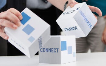 Auf dem Bild sind mehrere Würfel zu sehen. Die Würfel tragen die Begriffe „CONNECT“, „INNOVATE“, „CHANGE“ und „LEARN“. Eine Person hält einen Würfel, während im Hintergrund weitere Personen sichtbar sind. Der Kontext deutet auf eine kreative oder strategische Diskussion hin. Es geht um dase lektronische Handelssysteme MACD.