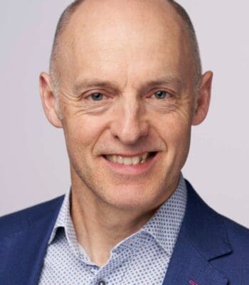Das Bild zeigt Jon O'Donnel, COO Acuity Knowledge Partners - einen Mann mit kahlem Kopf und lächelndem Gesichtsausdruck. Er trägt ein blaues Sakko über einem hellen, gepunkteten Hemd. Der Hintergrund ist neutral und hell. Der Mann wirkt freundlich und professionell, was auf einen geschäftlichen Kontext hindeutet.