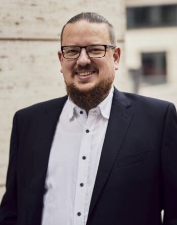 Dauerbrenner Mainframe - Aus der Praxis: der Weg in die Cloud, ein Beitrag von Boris Bathow und Philipp Koch, Partner bei Senacor Technologies. Auf dem Bild ist Philipp Koch mit Brille und einem gepflegten Bart zu sehen. Er trägt einen schwarzen Anzug und ein weißes Hemd. Der Hintergrund zeigt eine unscharfe, moderne Gebäudeansicht. Der Mann lächelt freundlich in die Kamera und vermittelt einen professionellen Eindruck.
