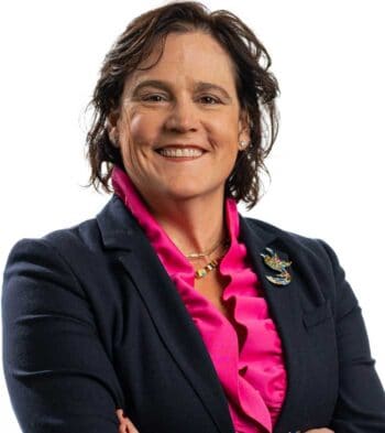 Digital transformation Lead, Global Financial Services, Red Hat - Frau mit kurzen, lockigen Haaren. Sie trägt ein dunkelblaues Sakko und eine pinke Bluse. An ihrem Sakko ist ein Abzeichen befestigt. Der Hintergrund ist einfarbig weiß, was den Fokus auf die Person lenkt.