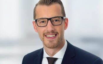 Das Bild zeigt einen Mann mit Brille, der in einem Anzug gekleidet ist. Er lächelt freundlich und steht vor einem unscharfen, hellen Hintergrund. Der Kontext deutet auf ein professionelles Umfeld hin, möglicherweise für ein Bewerbungsfoto oder ein Unternehmensporträt.