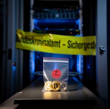 Das Bild zeigt eine durchsichtige Plastiktüte mit mehreren Kryptowährungen, darunter Bitcoin, Litecoin und Dash, die auf einem Tisch vor einer Serverinfrastruktur platziert ist. Im Hintergrund ist ein gelbes Banner mit der Aufschrift „Bundeskriminalamt – Sichergest.“ zu sehen. Diese Sicherstellung steht im Zusammenhang mit der Abschaltung des Krypto-Swapping-Dienstes „eXch“ durch das BKA und die ZIT.