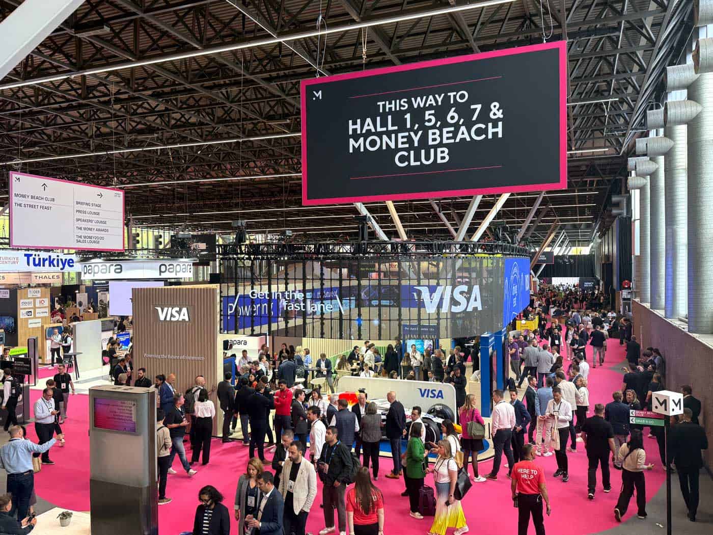Die Veranstaltung Money20/20 präsentiert eine lebhafte Messe mit zahlreichen Ausstellern und Besuchern. Auffällige Beschilderungen weisen den Weg zu verschiedenen Hallen und dem Money Beach Club. Die Atmosphäre ist geschäftig und fokussiert auf Integration und technische Lösungen.