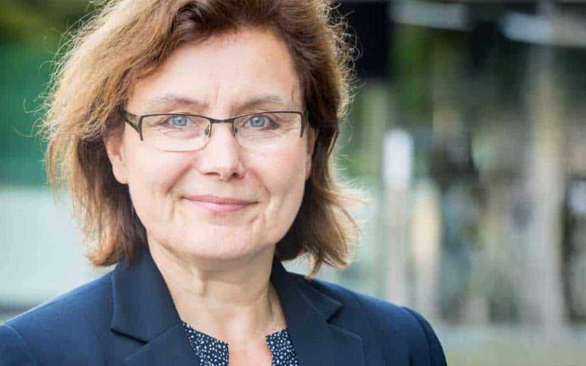 Eine Person mit braunen, lockigen Haaren und Brille lächelt in die Kamera. Sie trägt einen dunklen Blazer und ein gemustertes Oberteil. Der Hintergrund ist unscharf, was den Fokus auf die Person lenkt. Das Bild könnte im Kontext des Zahlungsverkehrs