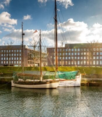 Im Vordergrund ist ein Boot im Wasser zu sehen, während im Hintergrund moderne Gebäude der Investitionsbank Schleswig-Holstein sichtbar sind. Die Szenerie wird von einem klaren Himmel und sanften Wolken begleitet.
